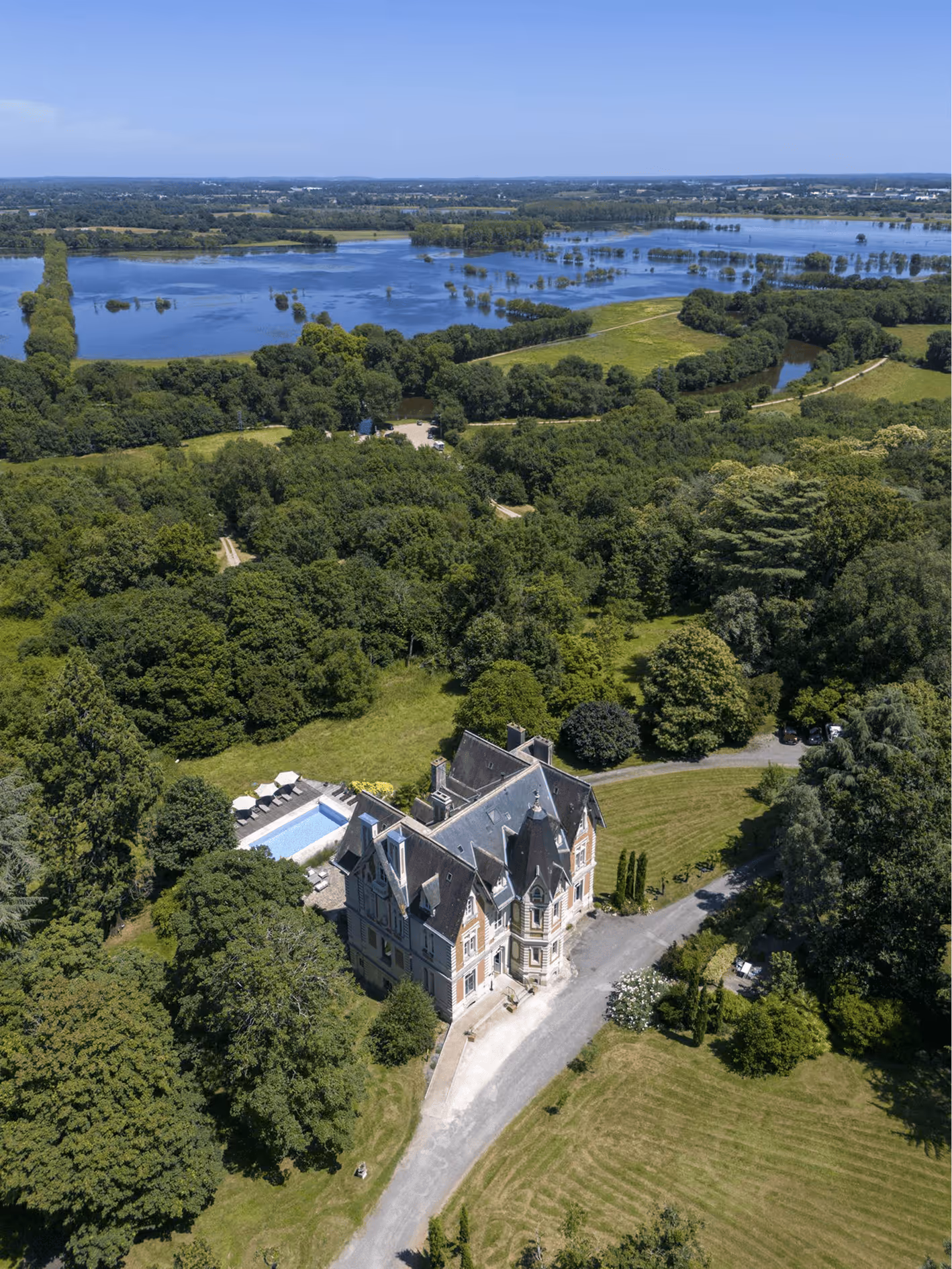 Vue aérienne d'un château entouré d'arbres avec une piscine et un paysage d'eau en arrière-plan.