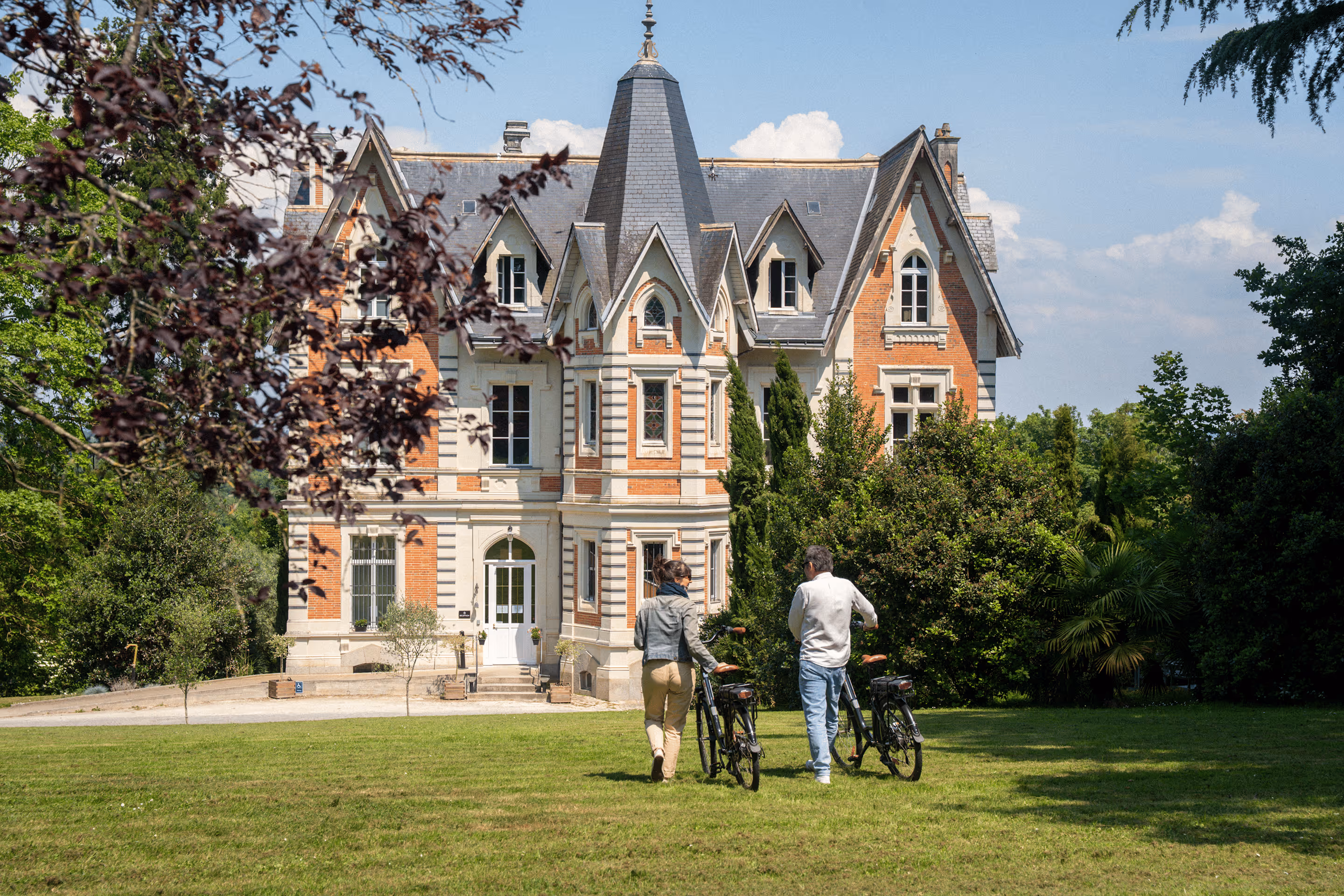 Deux personnes avec des vélos marchent vers un grand manoir victorien entouré d'arbres.