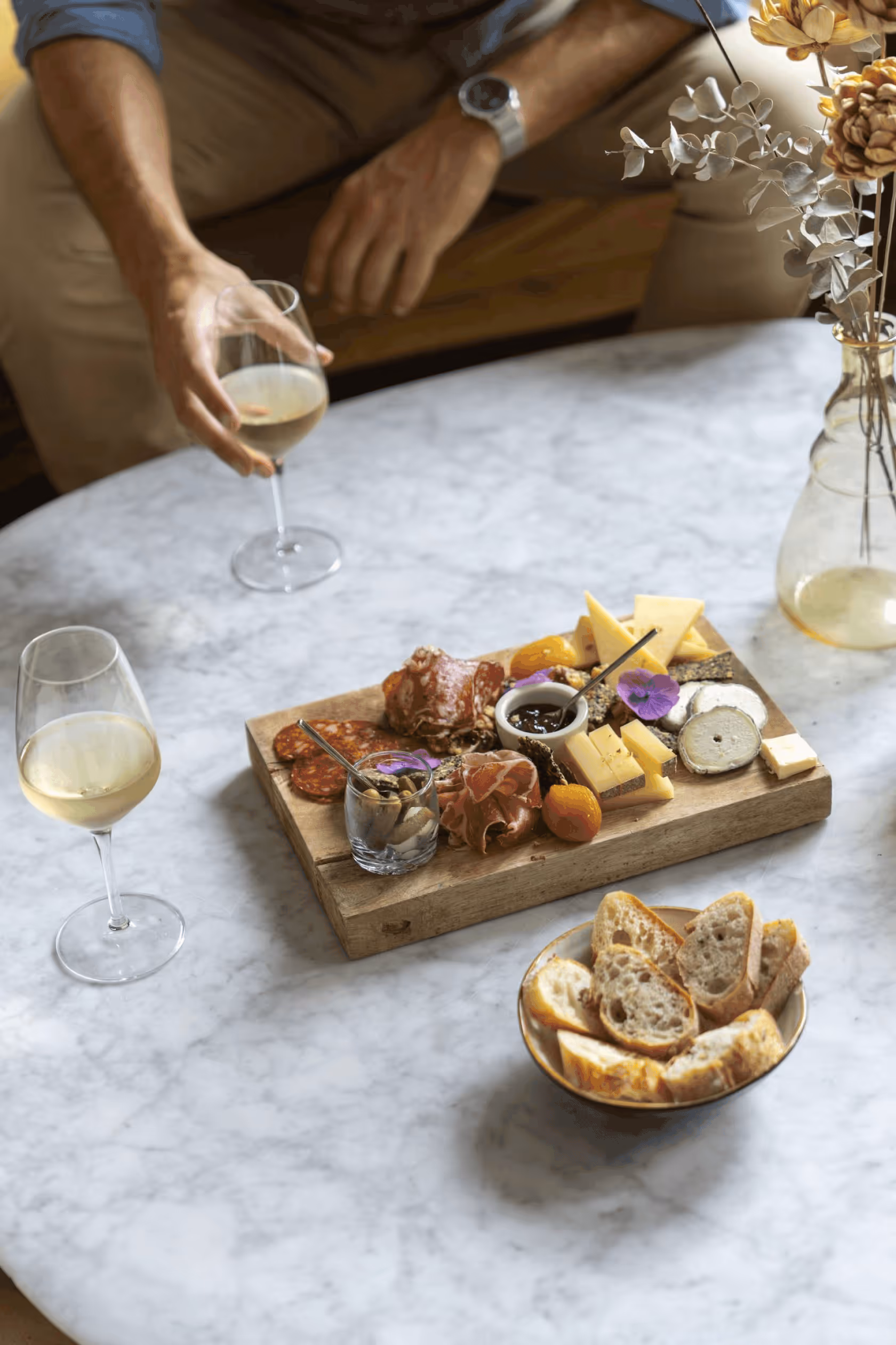 Person holding a glass of white wine next to a table with cheese, charcuterie, bread, and wine glasses.