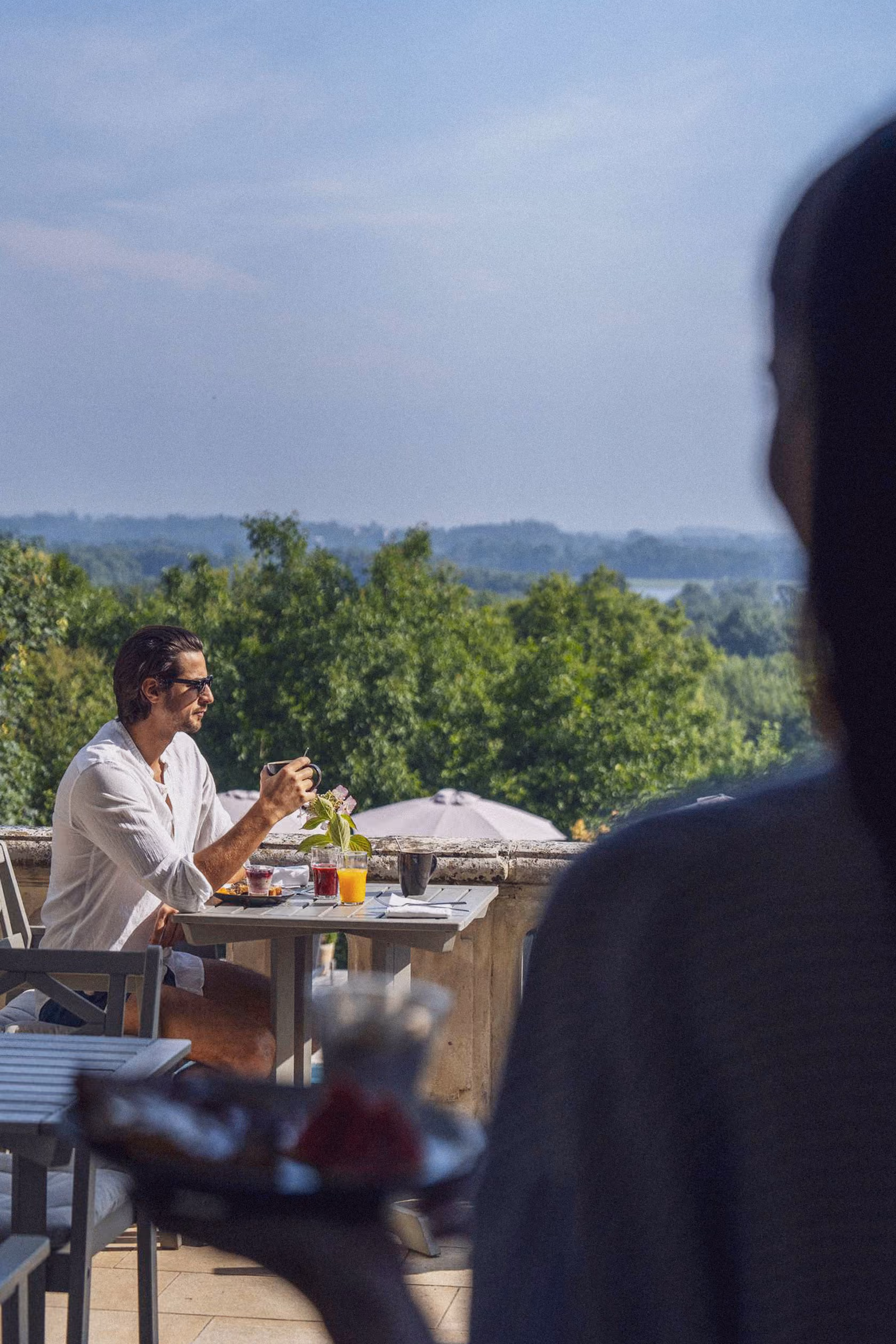 Un homme portant une chemise blanche et des lunettes de soleil est assis à une table en terrasse avec une tasse à la main, devant un paysage verdoyant sous un ciel dégagé.