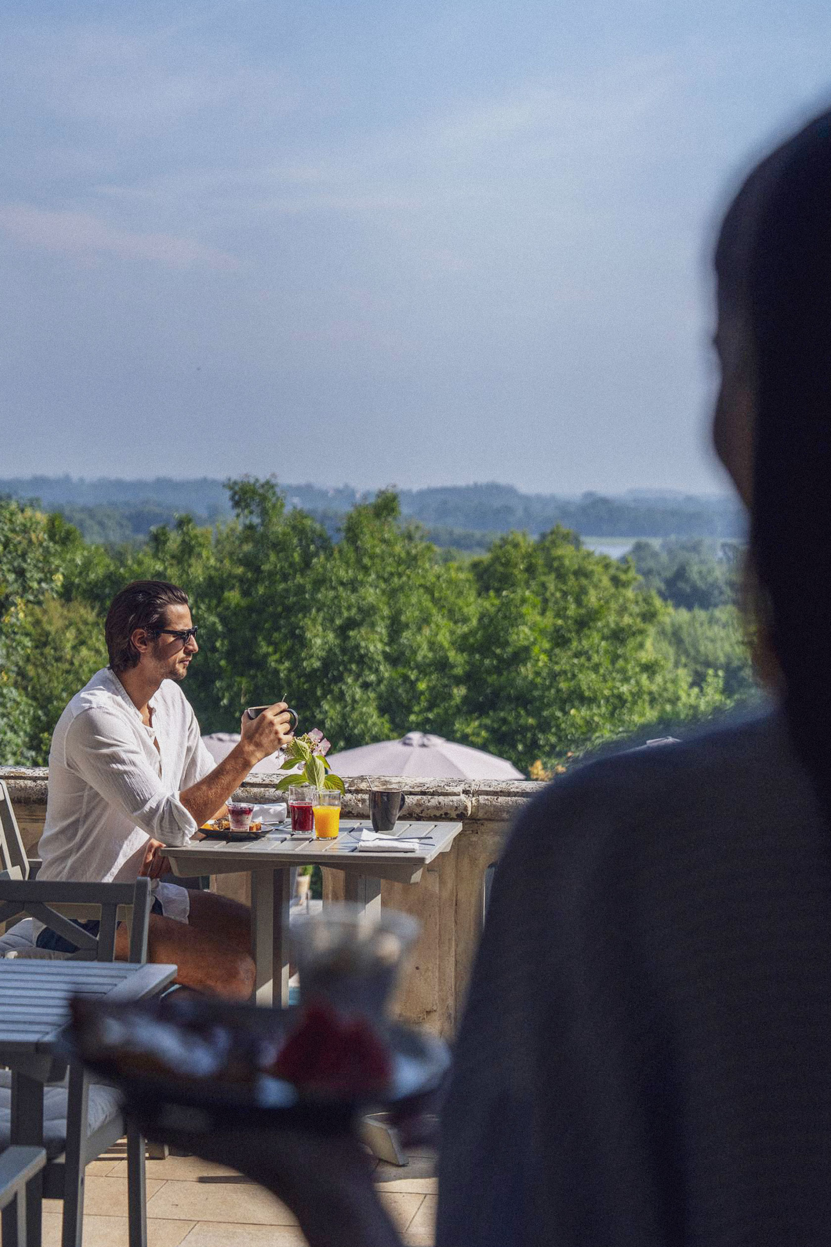 Un client prend son petit déjeuner au soleil sur la terrasse du château, rejoint par sa compagne qui rentre en amorce dans l'image.