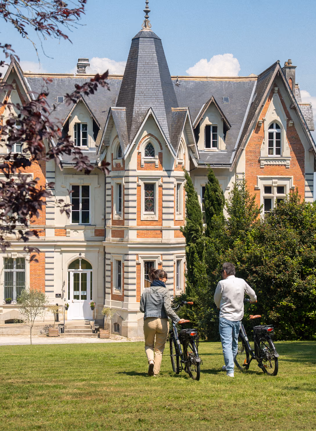 Deux personnes avec des vélos marchent vers un grand manoir victorien entouré d'arbres.