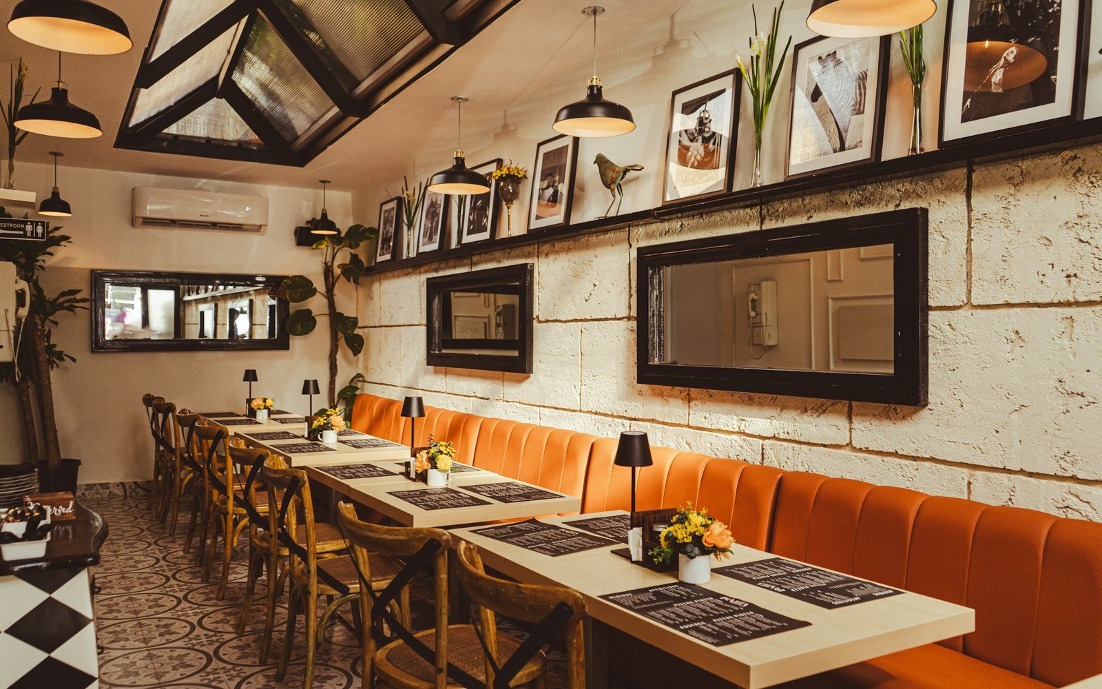 A wide photo of a restaurant dining room with an orange banquette and framed pictures on the wall