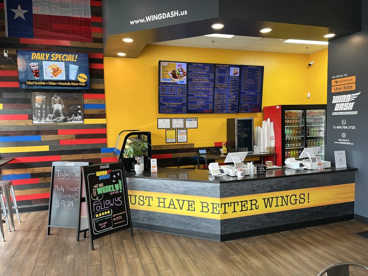 The Wing Dash counter in Dallas, with four menu boards on a yellow wall and a Daily Special screen