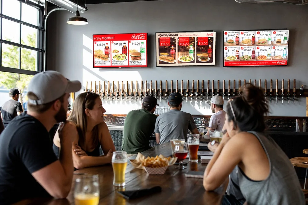 Illustration of a brewery taproom: three TVs over a long row of taps show real Just a Menu combo and lunch specials boards