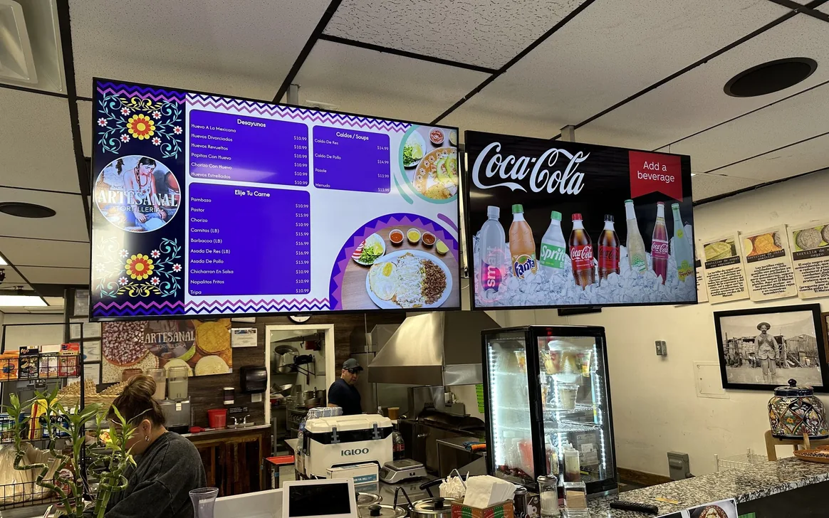 A purple menu board beside a Coca-Cola drinks board with bottled drinks on ice, over the counter at Artesanal Tortilleria