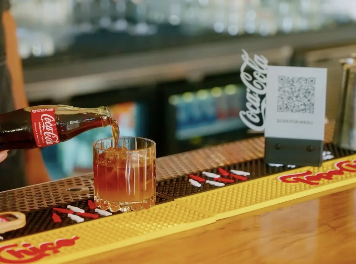 A bartender at Verbena in Austin pours a bottle of Coca-Cola over ice, next to a Scan for menu QR code stand on the bar