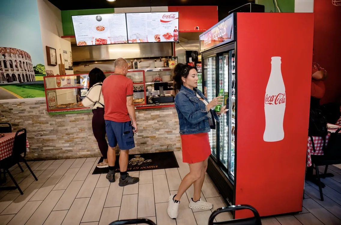 Guests ordering at the counter of Pizza Di Roma in Austin, under two menu boards, next to a drink cooler