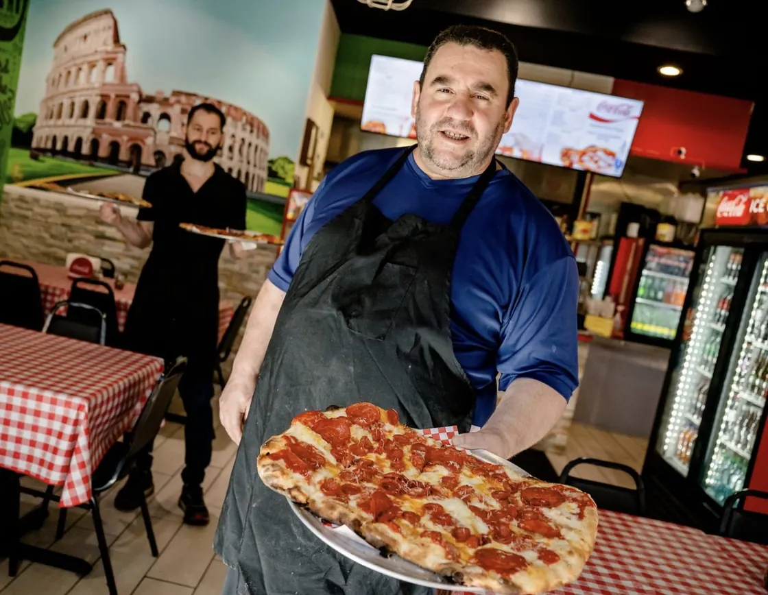 Tony, owner of Pizza Di Roma in Austin, holding a pepperoni pizza in his dining room, his menu boards on the wall behind