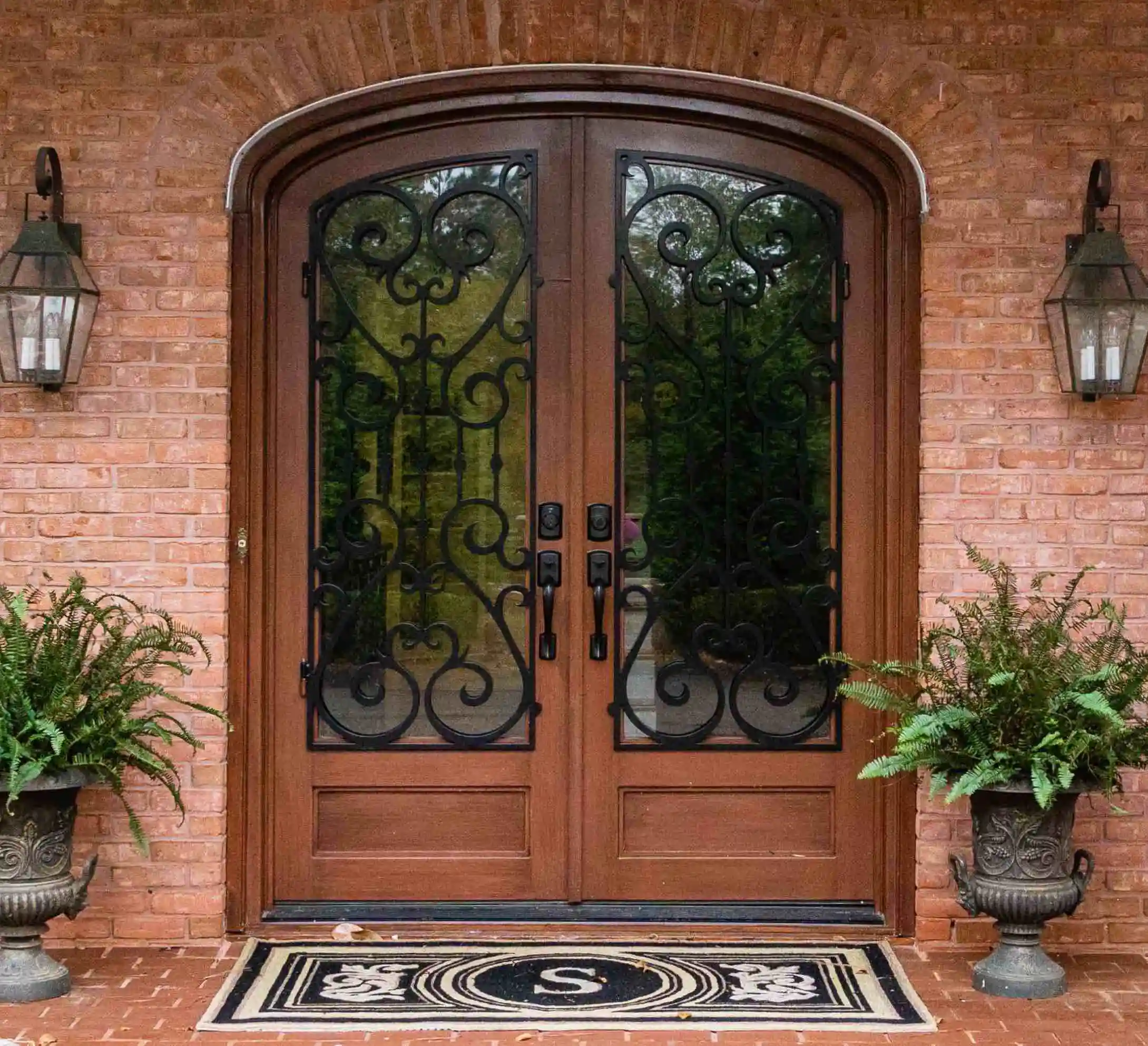 the front of a red brick home, in the middle there is a iron door made to look like dark wood with large panes of glass on each door with intricate black iron swirls on each. The door is slightly curved at the top