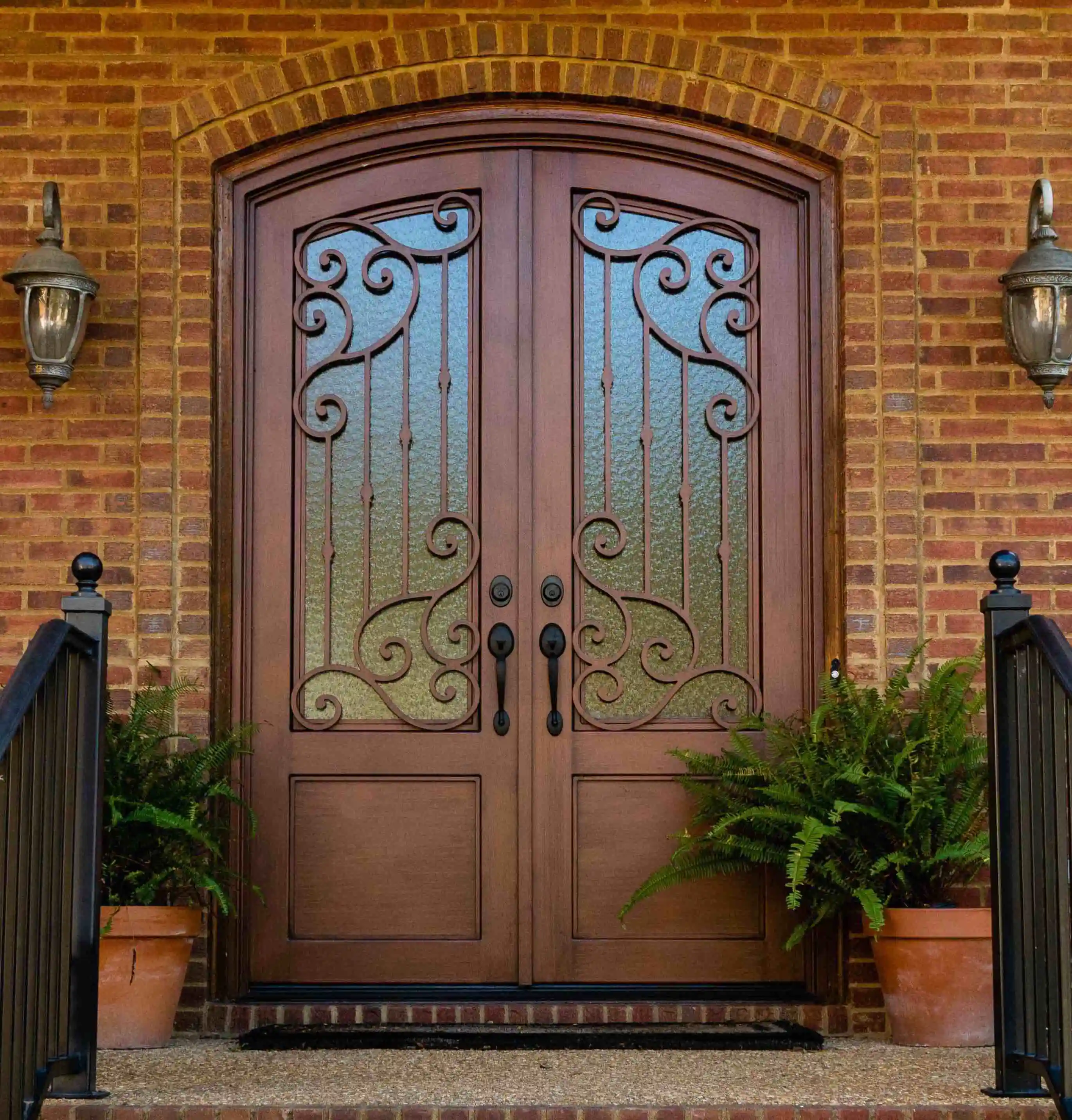 the front of a red brick home, in the middle there is a iron door made to look like dark wood with large panes of glass on each door with intricate black iron swirls on each