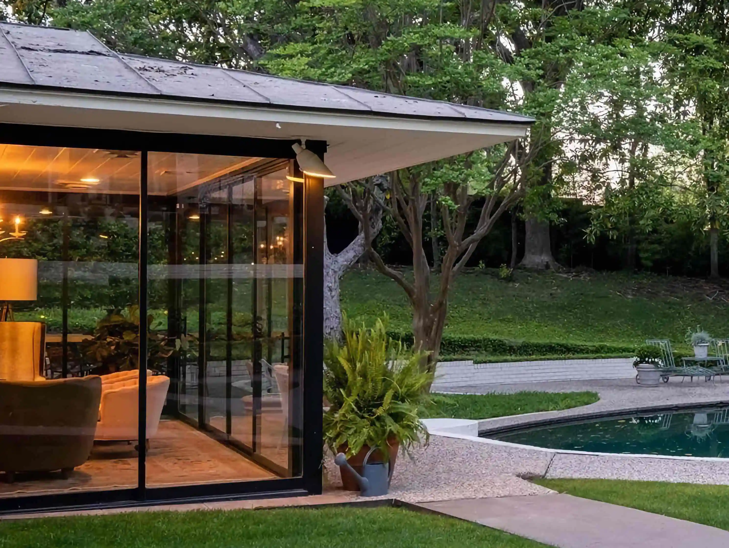the backyard of a home with the left side of the image being a large glass room of the house and the right side showing a pool with woods behind it
