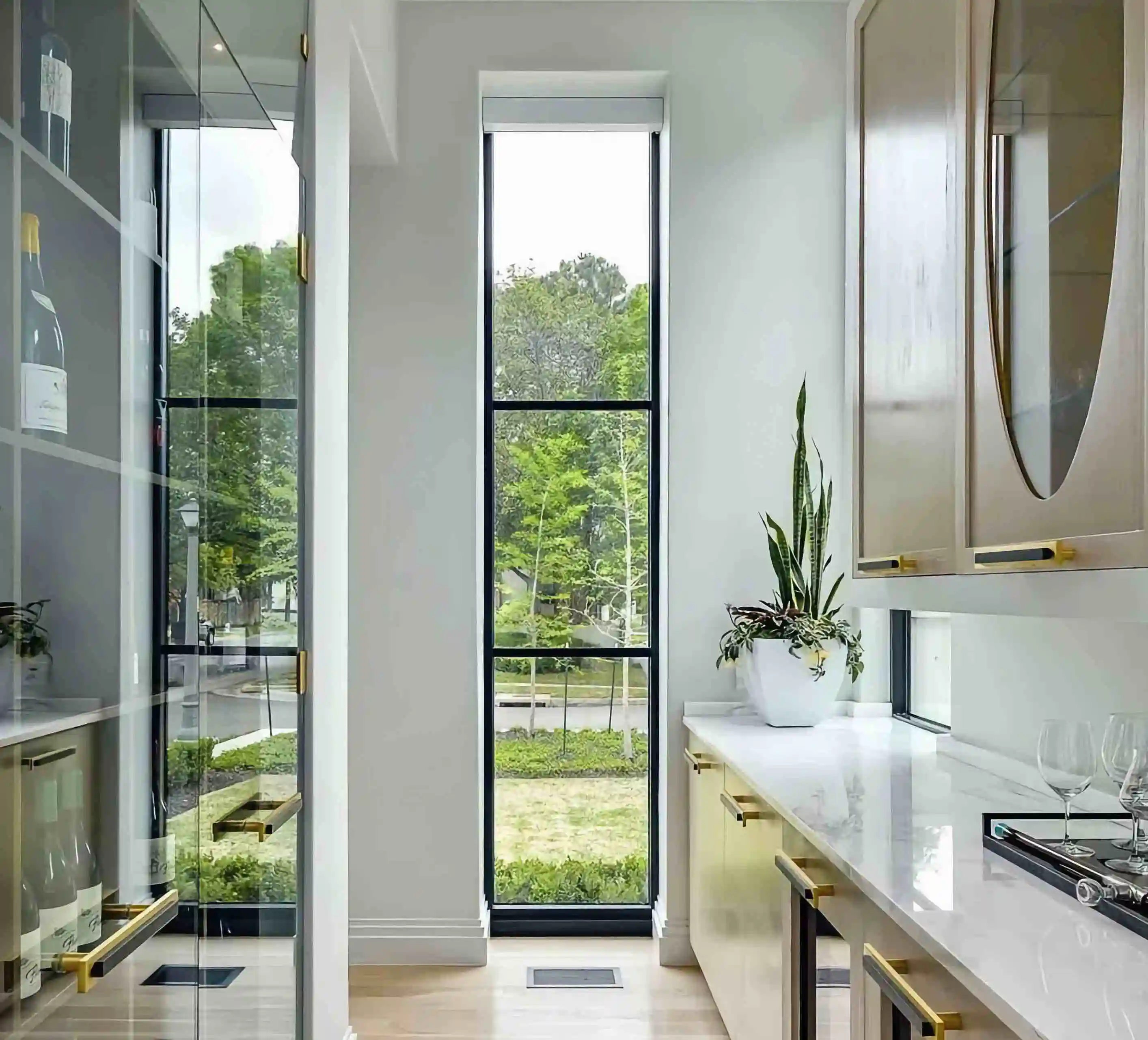 inside the bar/kitchen area of a home with a glass wall covered wine cellar on the left and white countertops to the right and a skinny floor to ceiling glass window in front at the end of the room  