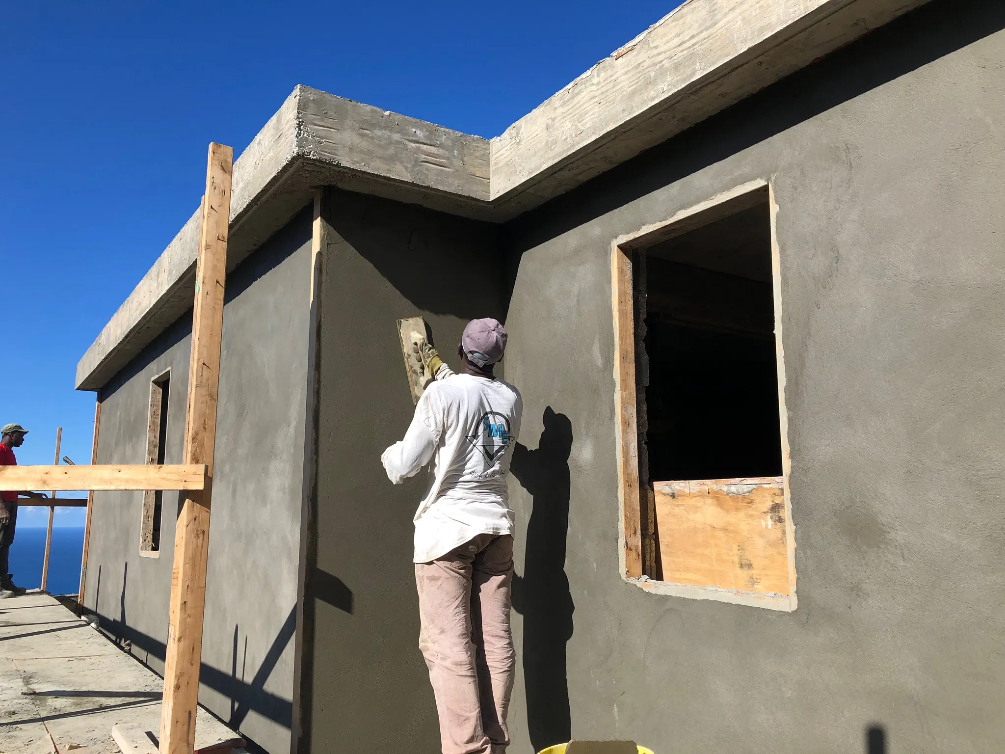 Two construction workers plastering the exterior walls of a concrete building under a clear blue sky.