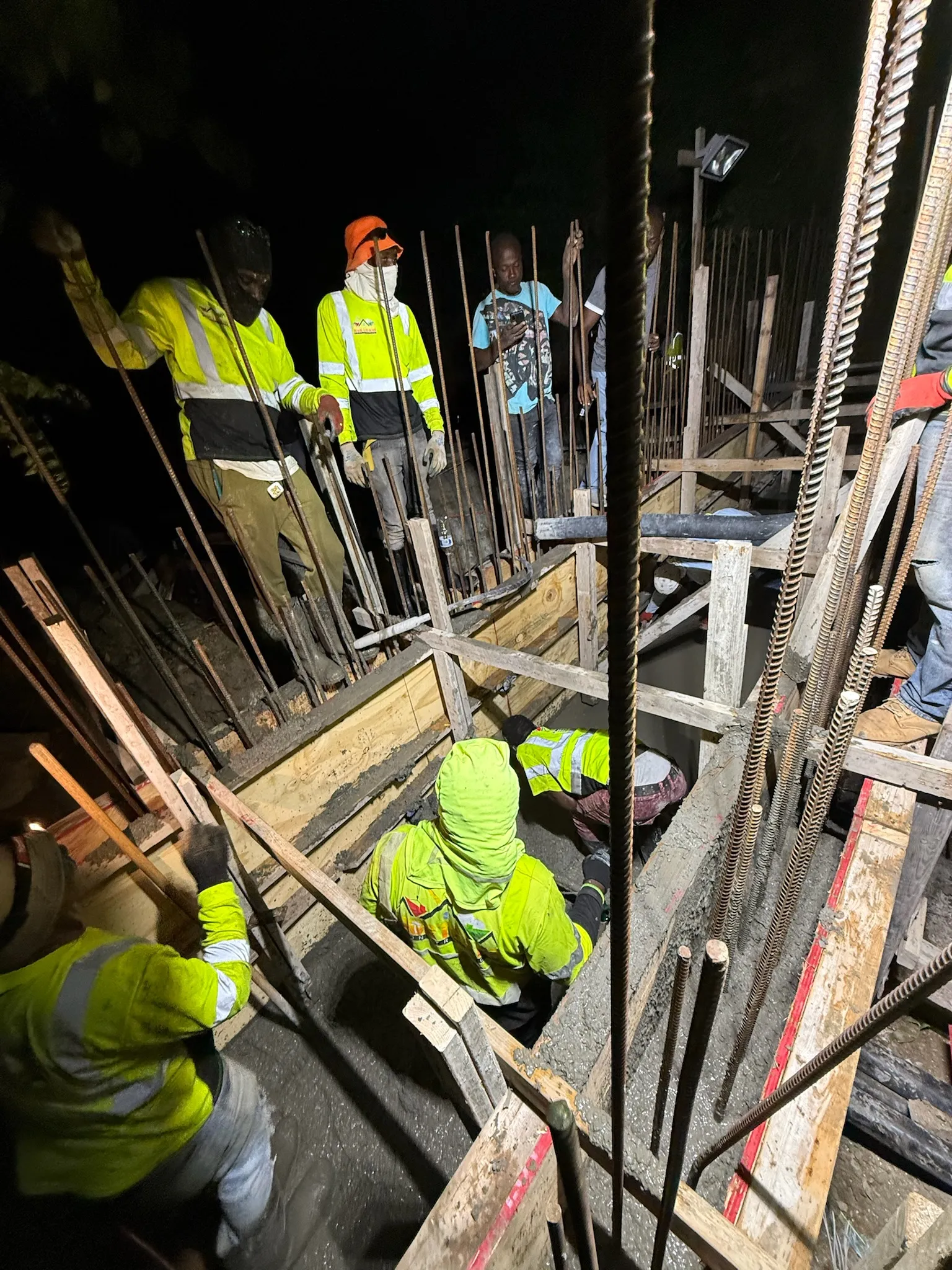 Construction workers in high-visibility clothing pouring and smoothing concrete at a nighttime construction site with metal rebar and wooden frames.