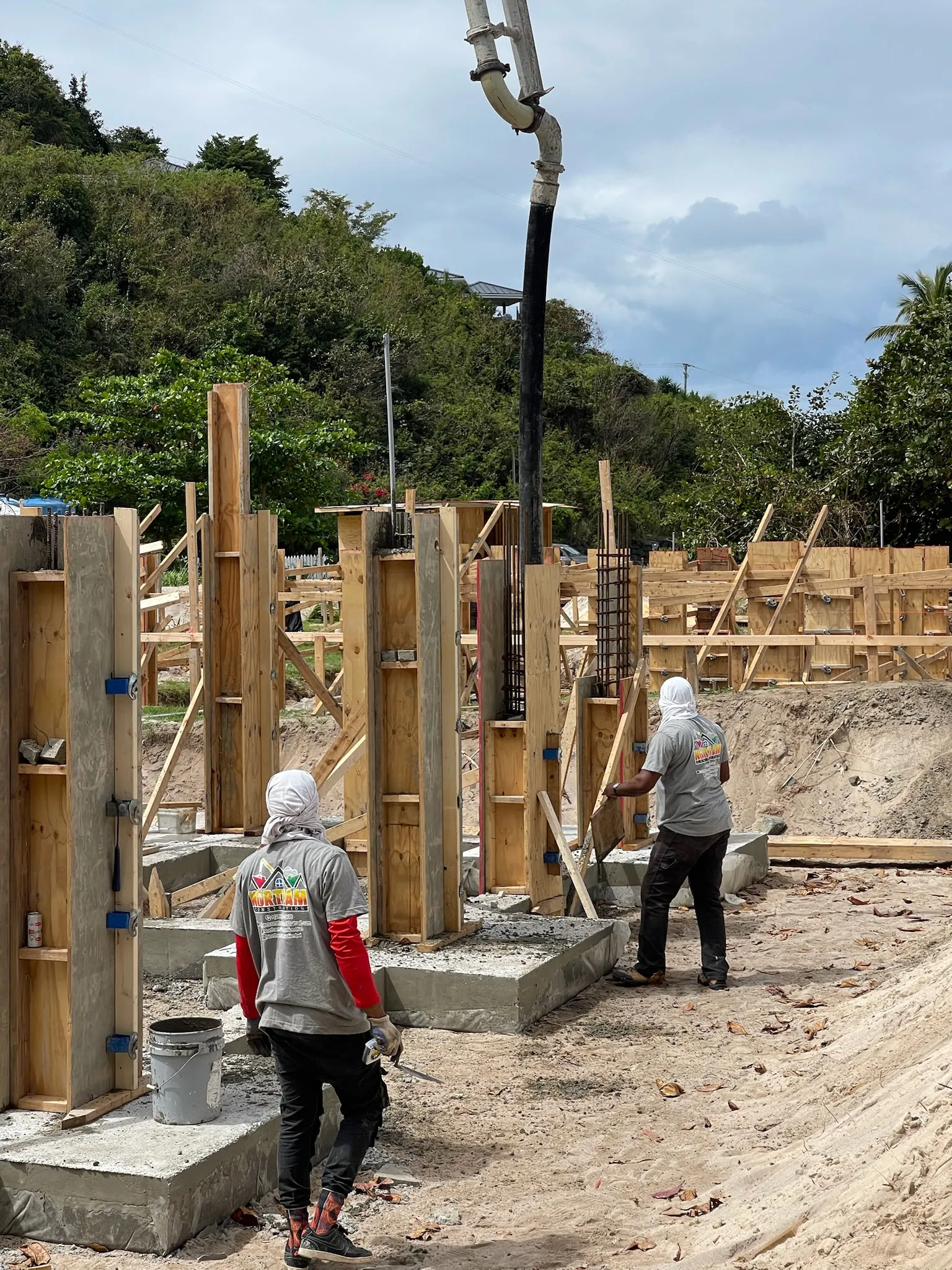 Two construction workers with face coverings pouring concrete into wooden formwork for pillars at a construction site surrounded by greenery.