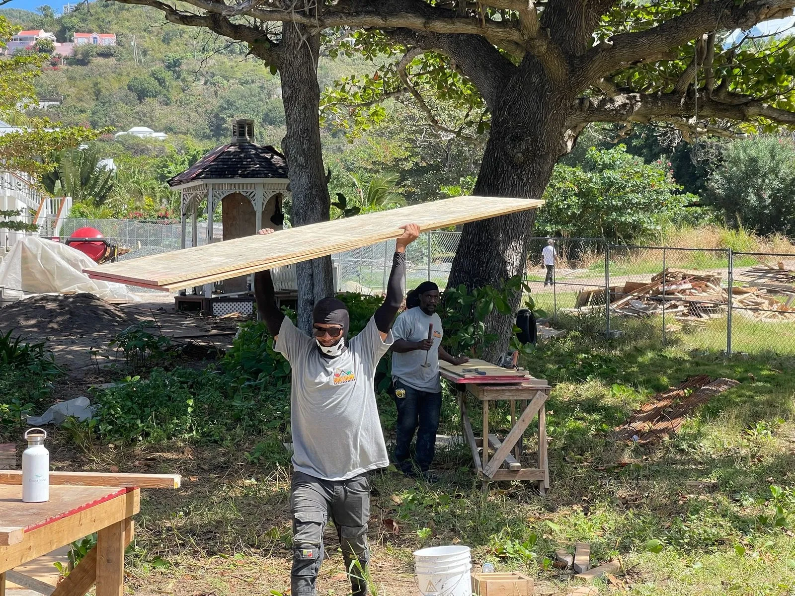 Construction workers outdoors carrying plywood sheets near trees and gazebo in a grassy area.