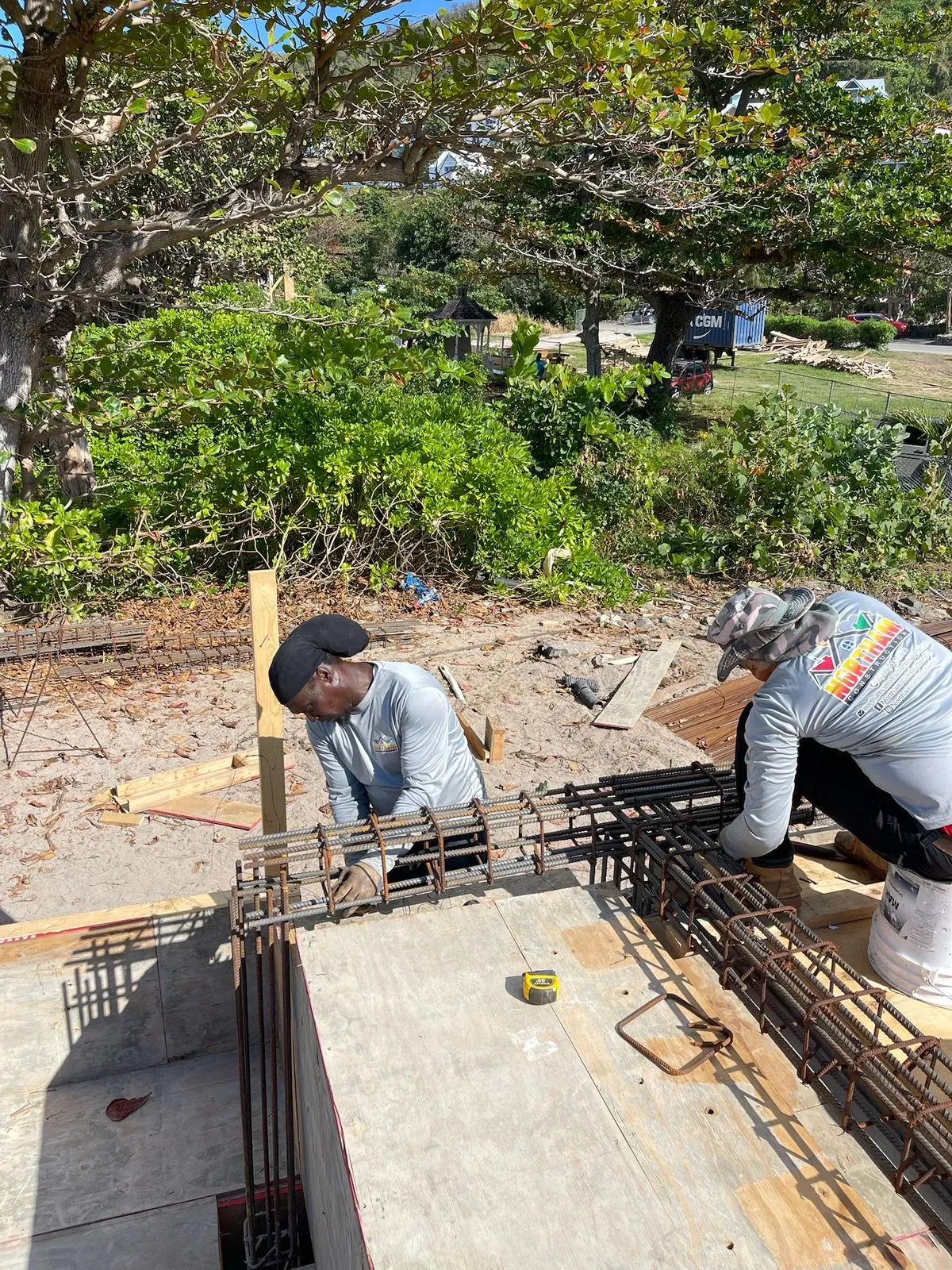 Two construction workers assembling steel rebar framework outdoors near green trees and bushes.