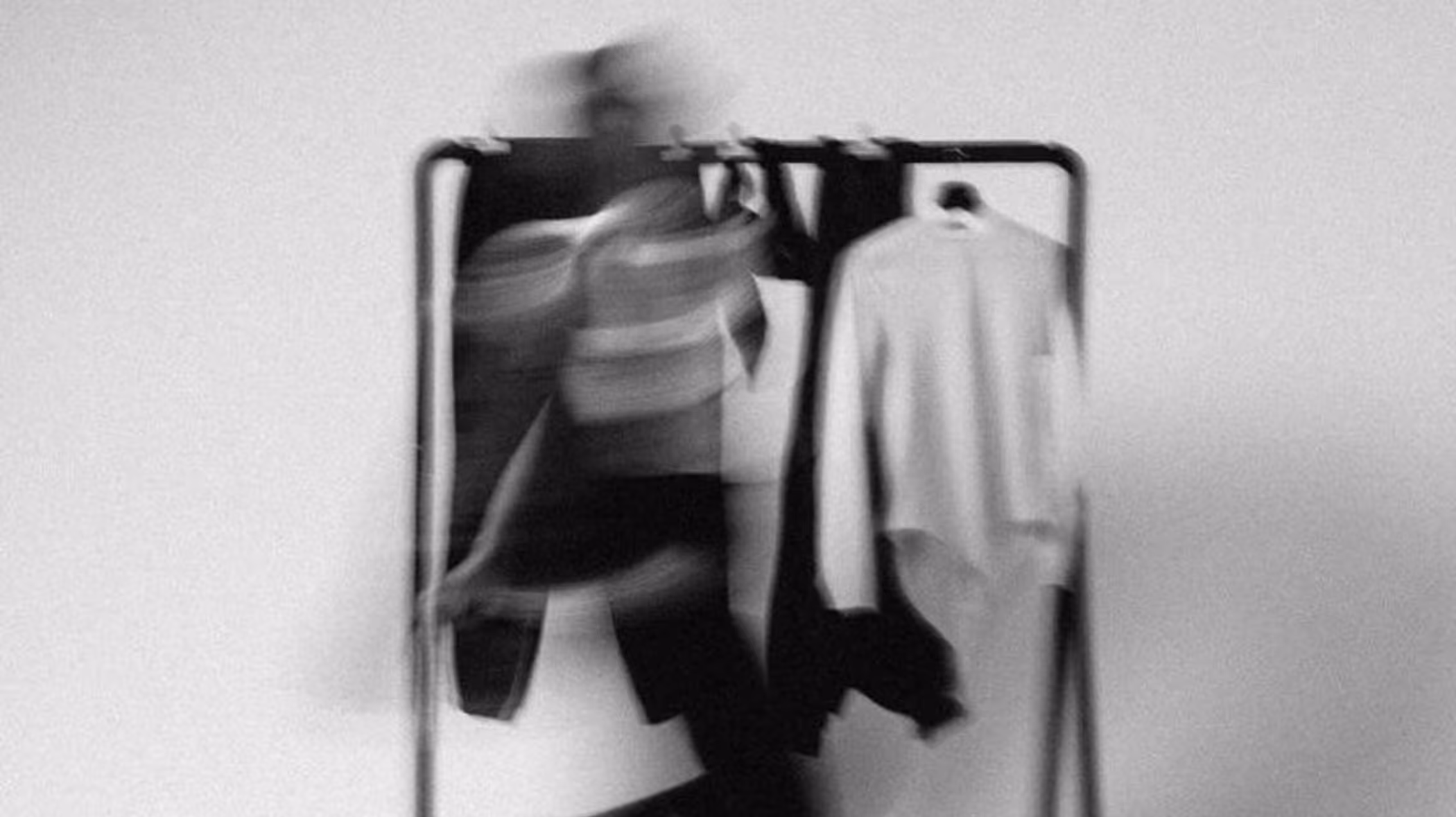 A blurred black-and-white image of a person walking past a clothing rack with shirts hanging on it.