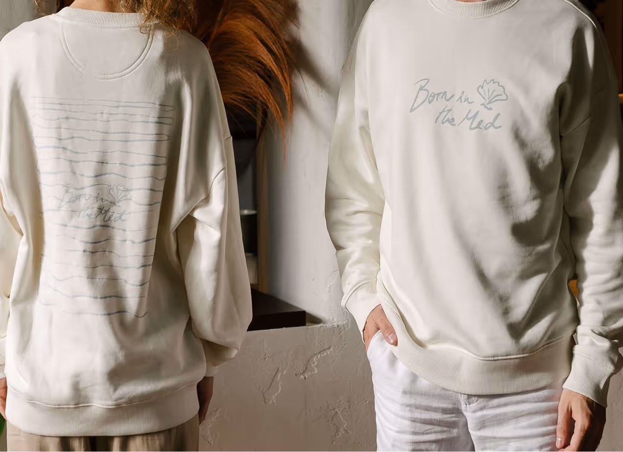 Two people wearing cream-colored sweatshirts with text and leaf designs; one shows the back with horizontal lines and text, and the other shows the front with the text "Born in the Med" and a leaf graphic.