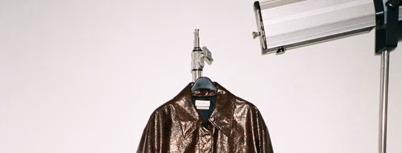 Un manteau en cuir marron brillant suspendu à un cintre noir devant un mur uni, avec une lampe de studio placée à droite.