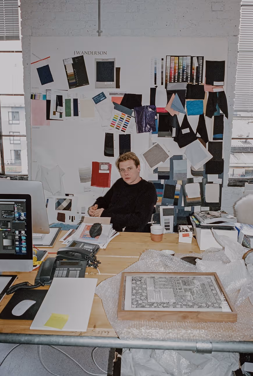 A man sitting at a wooden desk in an office surrounded by a computer, telephones, papers, and fabric swatches pinned to the wall.
