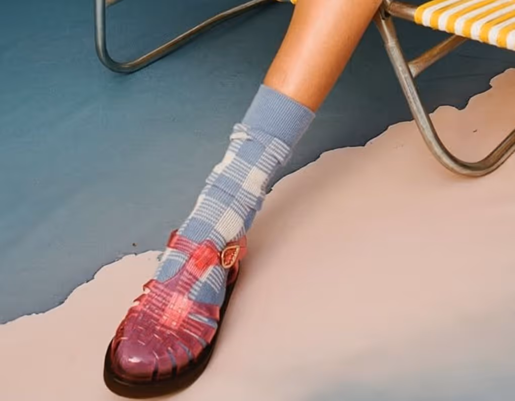 A leg wearing blue checkered socks and transparent pink sandals, resting on a yellow-and-white-striped chair.