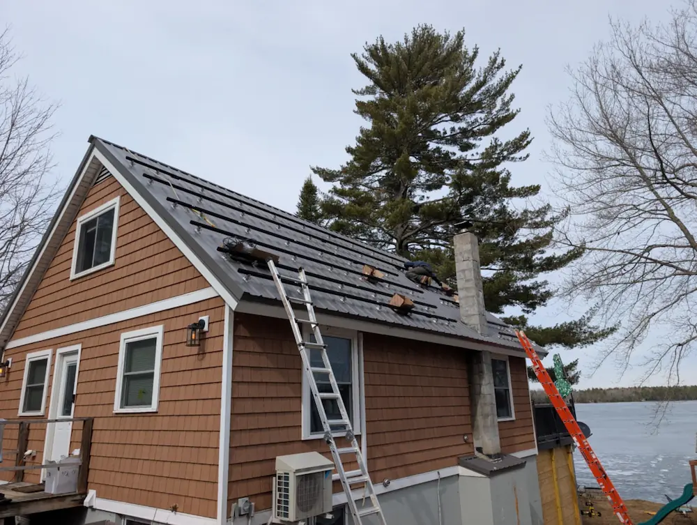 Workers installing dark shingles on the roof of a brown wooden house near a lake and trees.