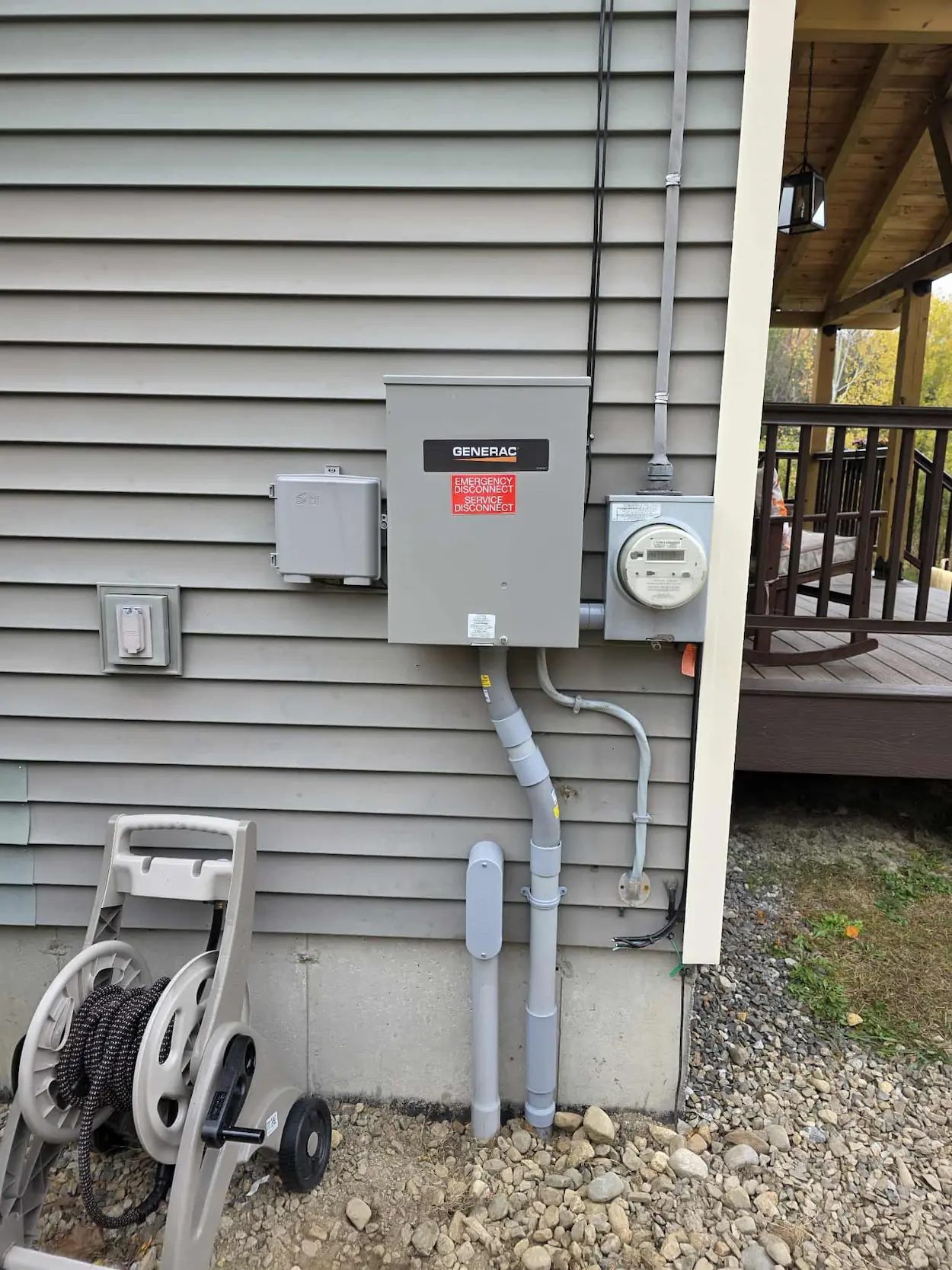 Gray electrical panel labeled Generac with emergency disconnect on house exterior with siding and gravel ground.