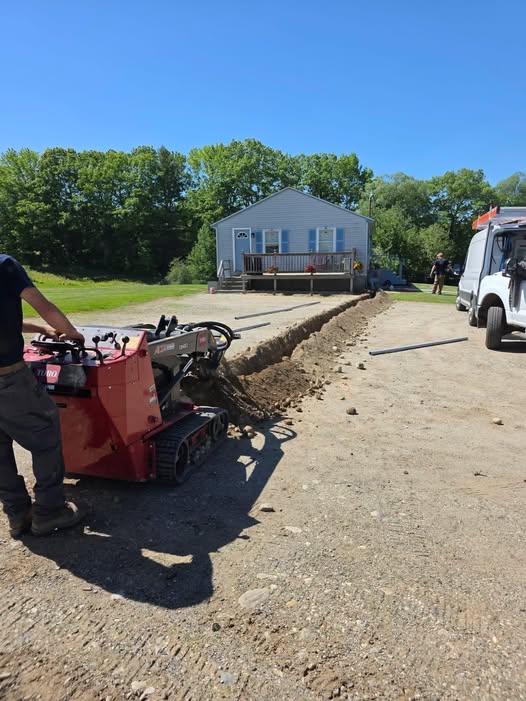 Worker using a red trencher machine to dig a trench on dirt driveway near a white house with trees.