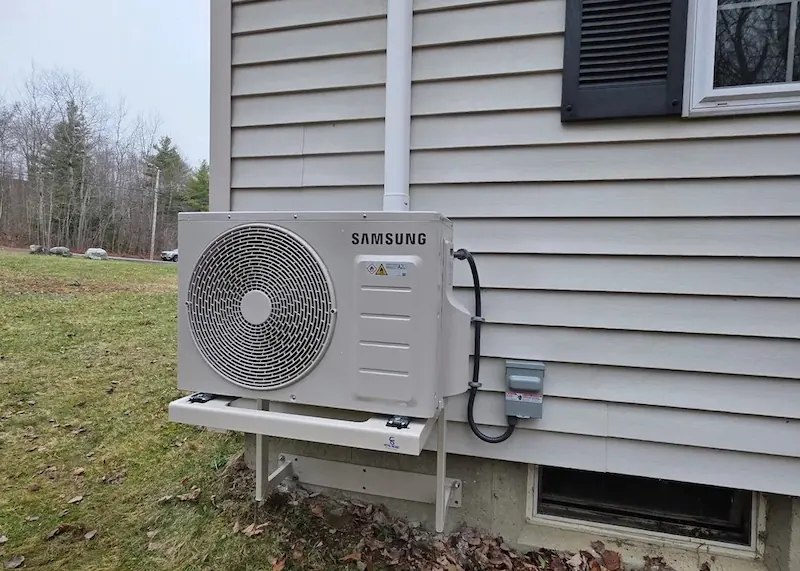 Samsung outdoor heat pump unit mounted on a beige house wall with a grassy lawn nearby.