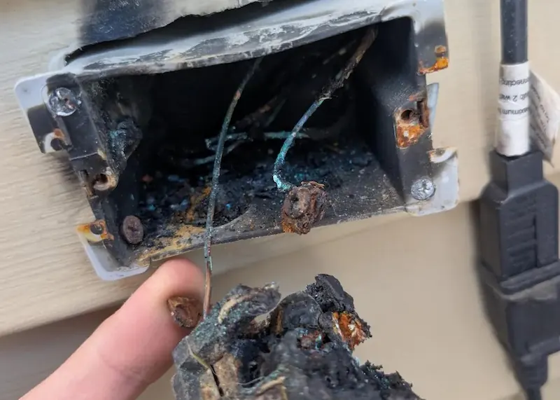 Close-up of a burnt and melted electrical outlet box with damaged wires and rusted screws.
