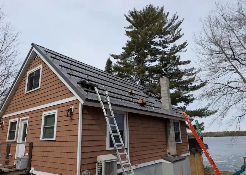 Workers installing solar panels on the roof of a lakeside wooden house with ladders leaning on it.