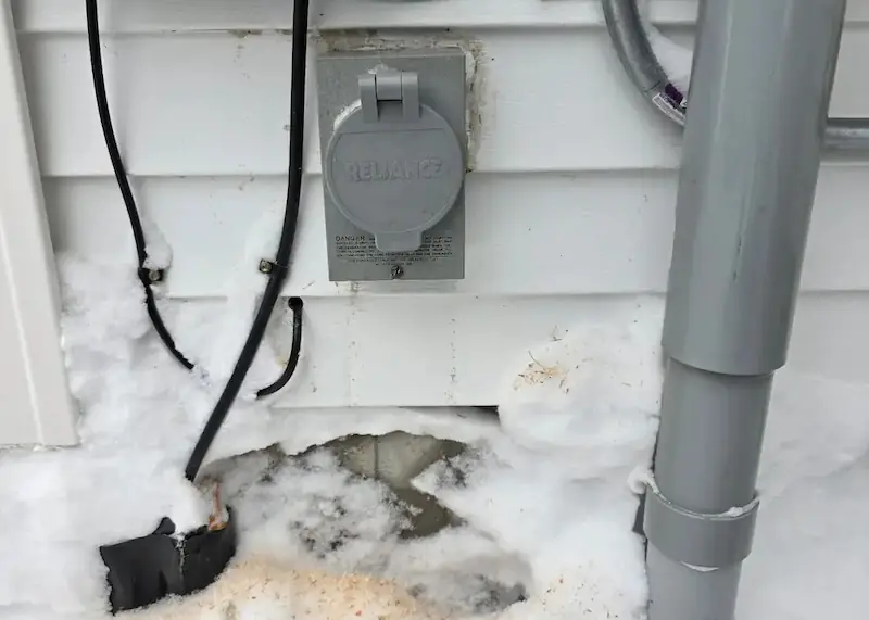 Electrical outlet with cover on exterior wall surrounded by snow and a large gray drainage pipe on the right.