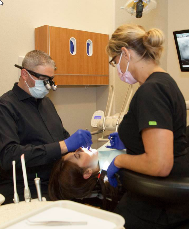 Dentist wearing magnifying loupes and a dental assistant working together to treat a patient during a dental procedure in a modern dental office. Both clinicians are wearing masks, gloves, and protective equipment while using dental instruments beside the reclining patient chair with cabinetry and treatment equipment visible in the background