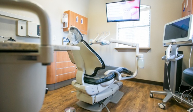 Modern dental treatment room featuring a reclining dental chair, mounted examination light, digital monitor, and advanced dental equipment with warm wood cabinetry and wood-style flooring creating a clean and comfortable patient environment
