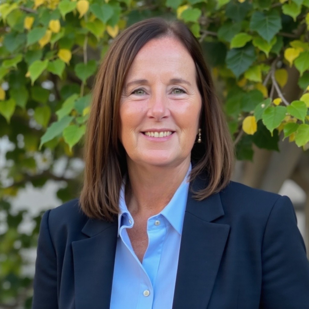 Smiling woman with shoulder-length brown hair wearing a blue shirt and dark blazer outside near green trees.