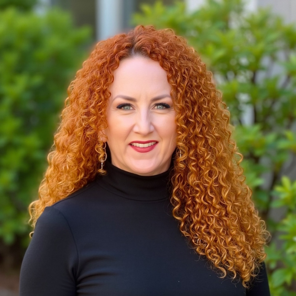 Smiling woman with long curly red hair wearing a black turtleneck, standing outdoors with greenery.