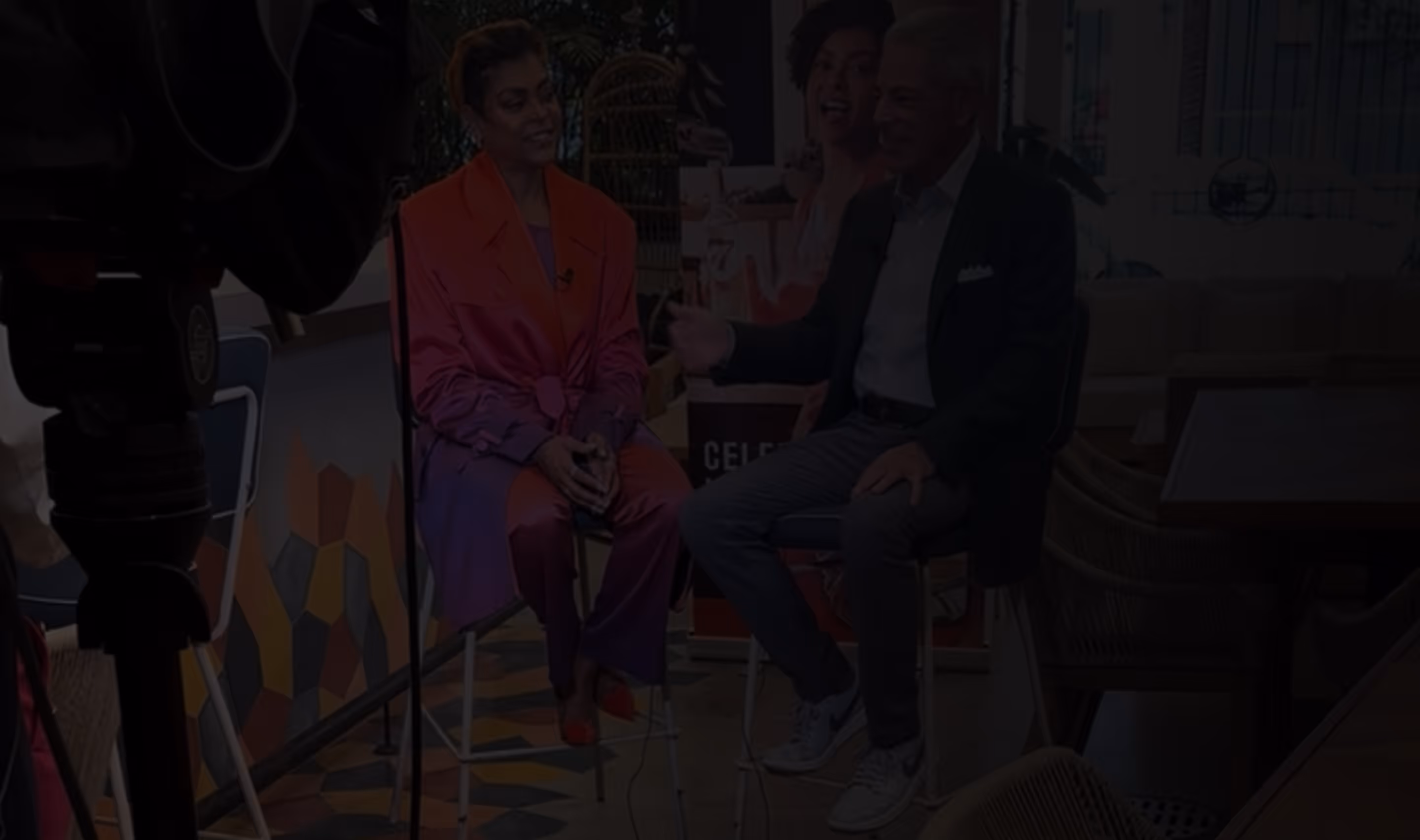 Two people sitting on high stools engaged in a conversation in a colorful indoor setting with a camera in the foreground.