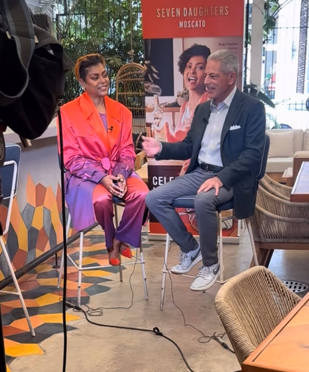 Two people sitting on stools having an interview in a colorful room with a Seven Daughters Moscato banner behind them.