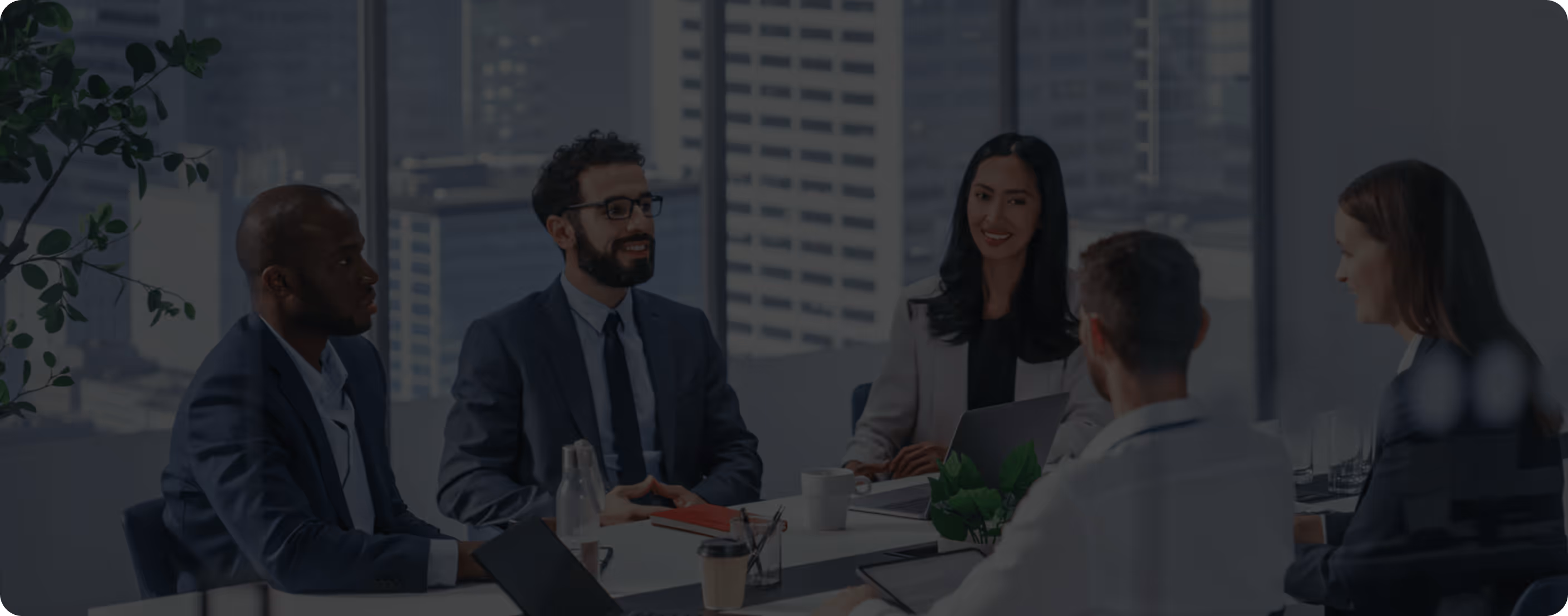 Five business professionals in suits sitting around a table in a modern office having a discussion.