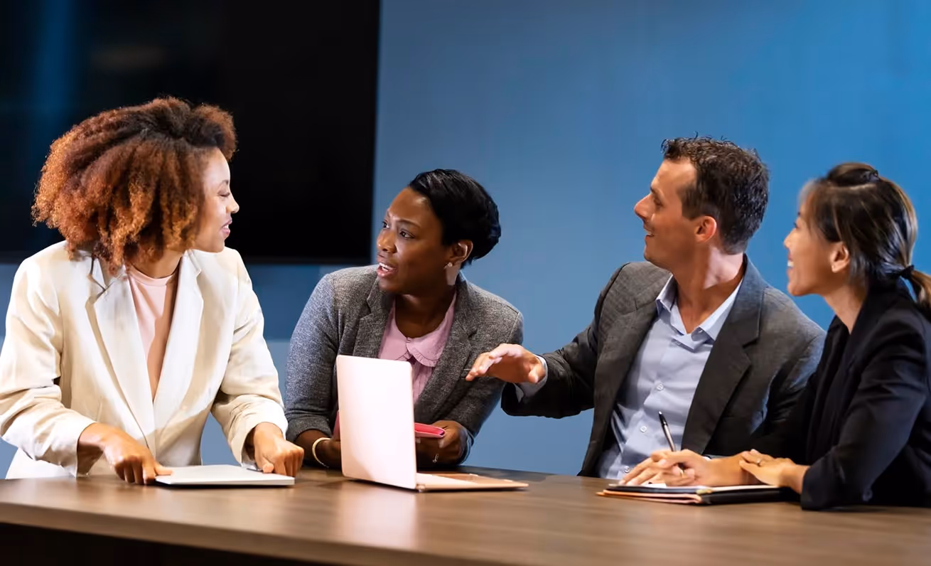 Four colleagues engaged in a discussion around a conference table with laptops and notebooks.