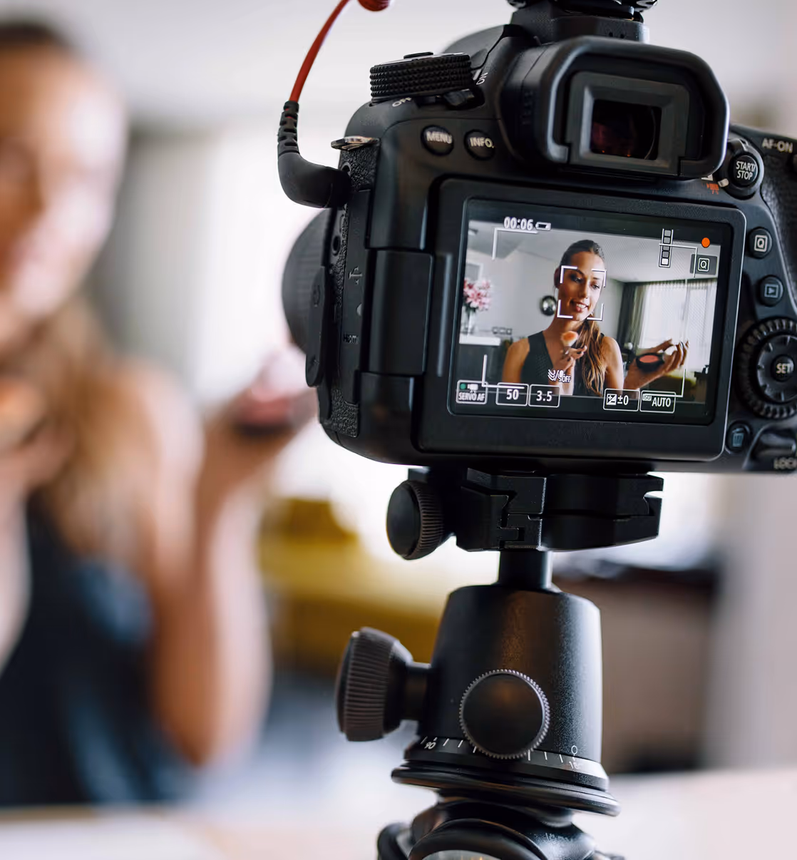 Digital camera on tripod recording a woman applying makeup indoors.
