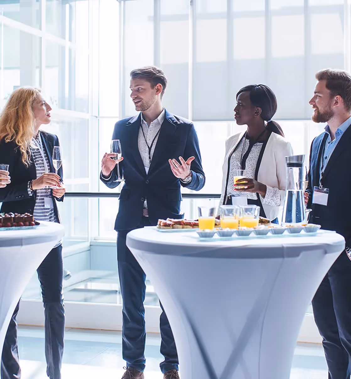 Four business professionals standing and conversing around cocktail tables with drinks and snacks in a bright modern room.
