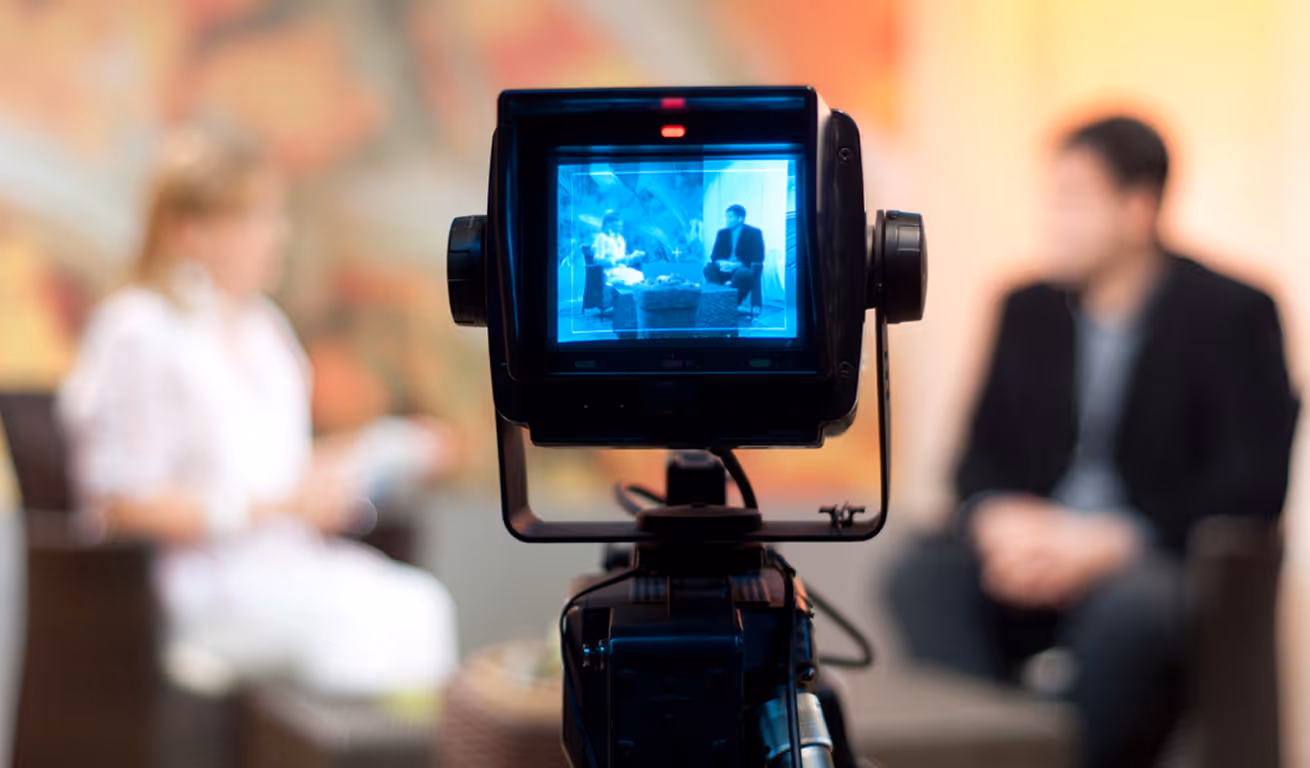 Television camera screen showing a man and woman having a studio interview on chairs.