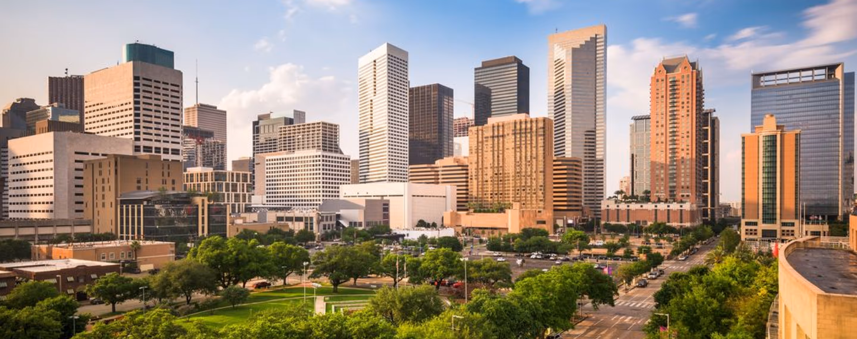 Downtown city skyline with tall buildings, green park, and tree-lined streets under a partly cloudy sky.