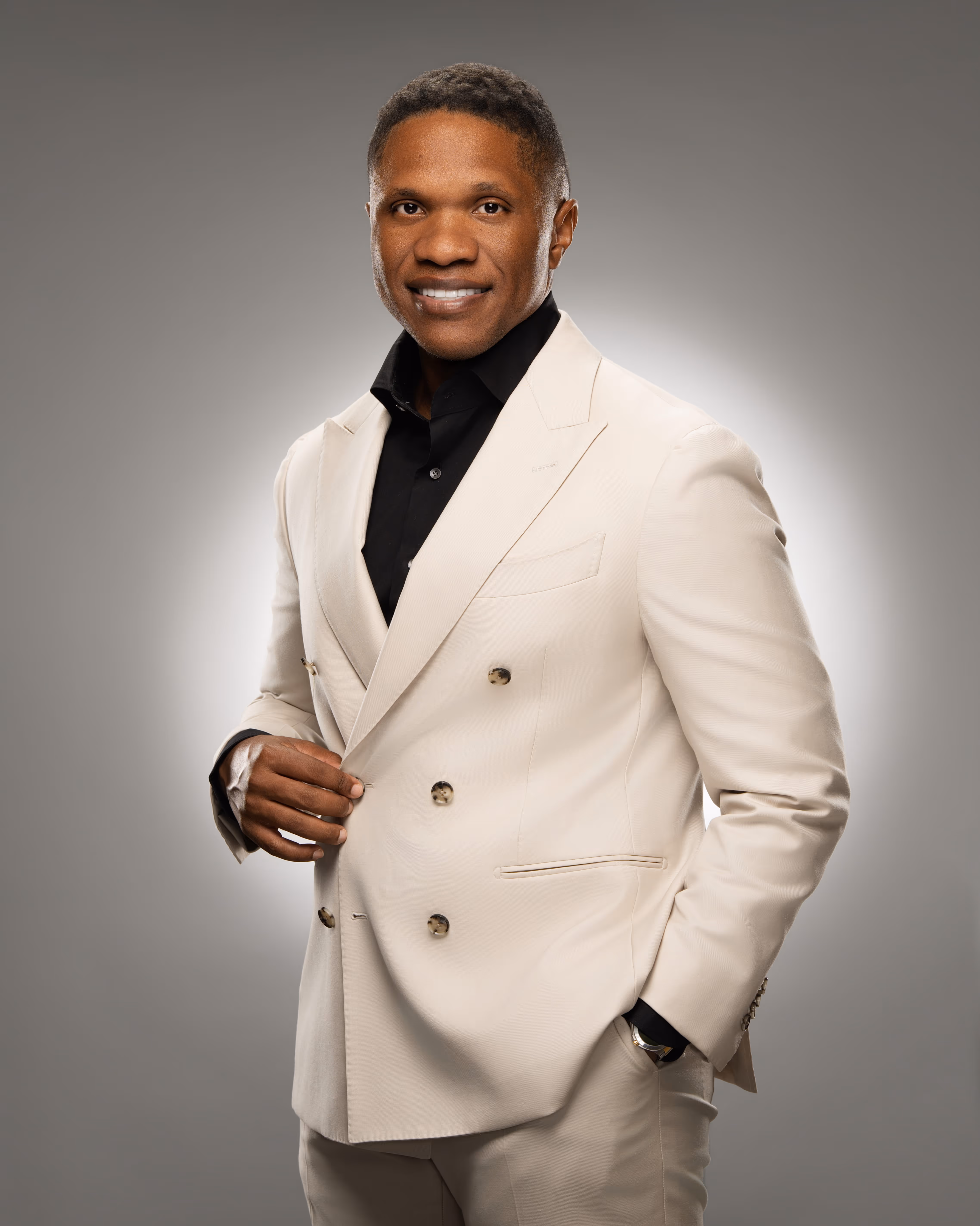 Man in a black suit sitting with hands clasped, smiling, with a piano in the background.