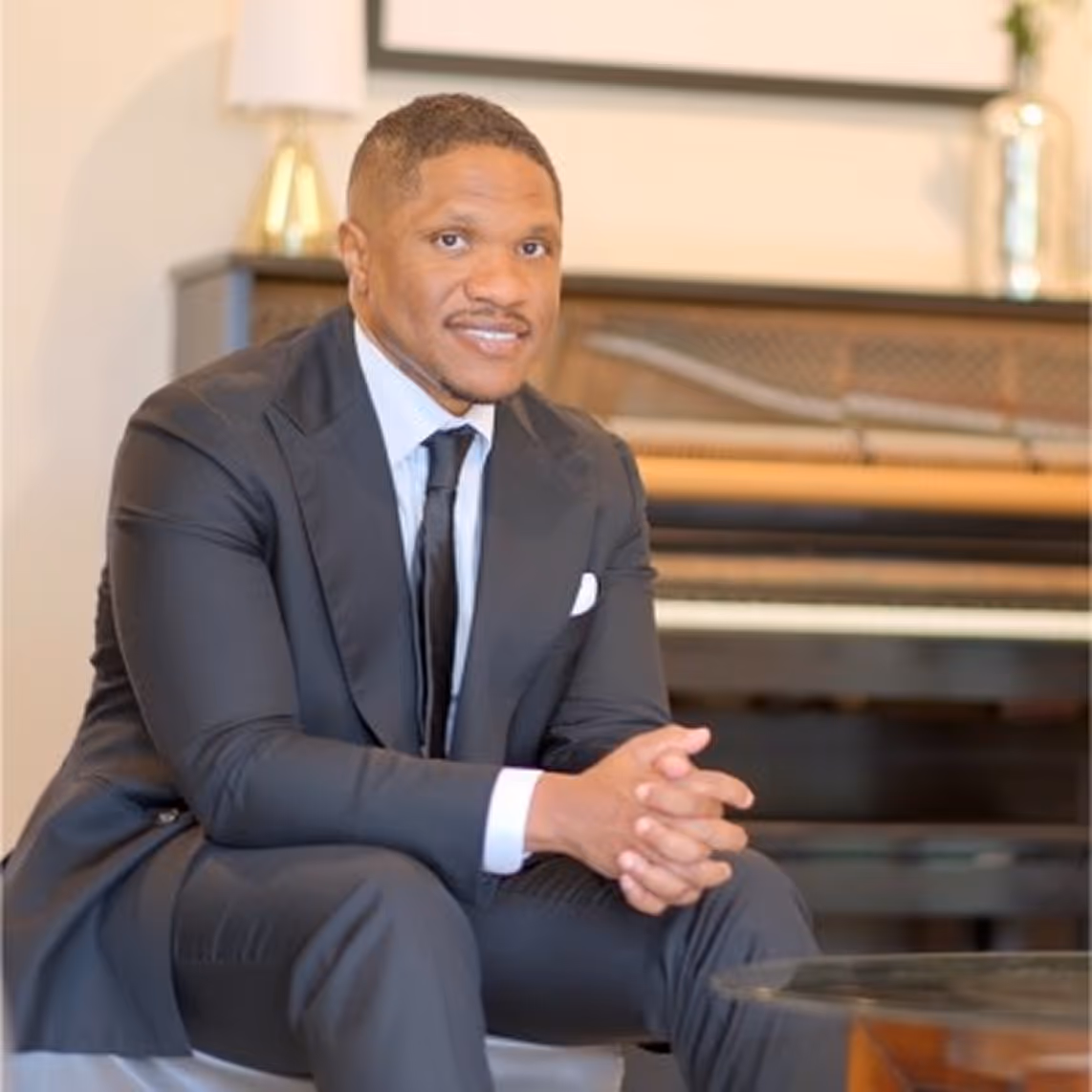 Man in a black suit sitting with hands clasped, smiling, with a piano in the background.
