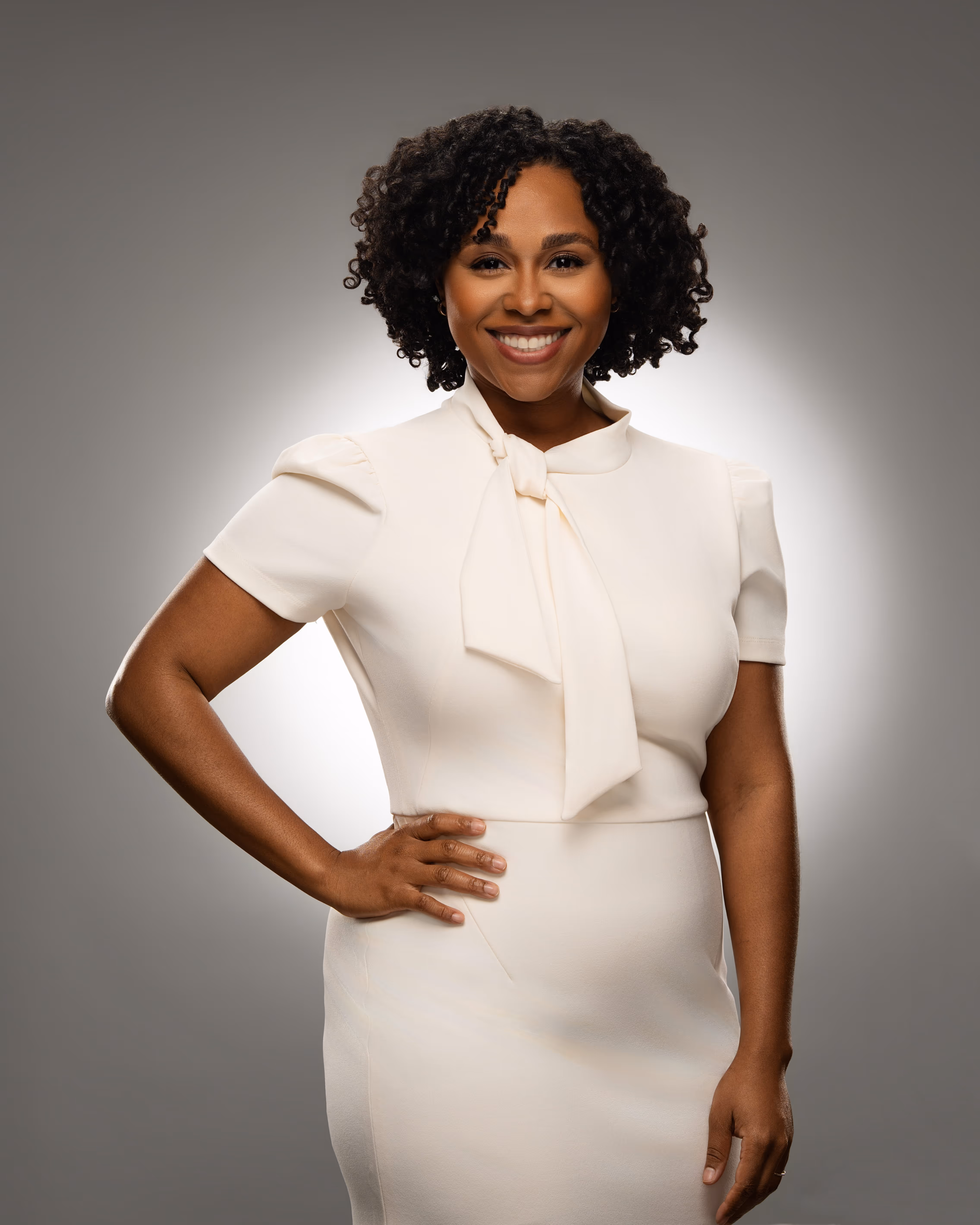 Smiling woman with curly hair leaning on a table near a laptop in a modern office.