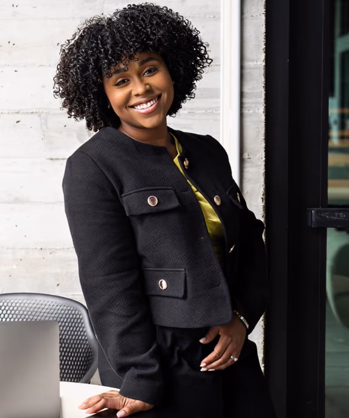 Smiling woman with curly hair leaning on a table near a laptop in a modern office.
